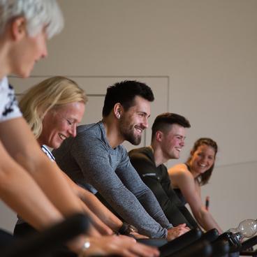 F&uuml;nf Personen auf Spinning-Bikes in einem Fitnessraum, l&auml;chelnd und konzentriert beim Training.