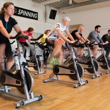 Gruppe von Personen beim Spinning in einem Fitnessstudio, mit Fahrr&auml;dern und Spiegeln im Hintergrund.