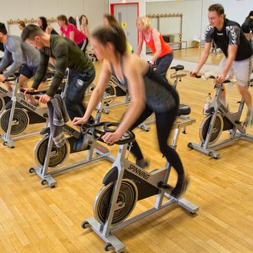 Gruppe von Personen auf Spinning-Bikes in einem Fitnessraum, mit Holzfu&szlig;boden und Spiegelwand im Hintergrund.