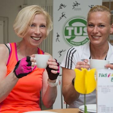 Zwei sportliche Frauen sitzen an einem Tisch und halten Tassen in der Hand, l&auml;chelnd vor einem TUS-Logohintergrund.