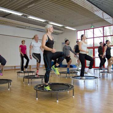 Gruppenfitness-Kurs mit Teilnehmern auf Trampolinen in einem hellen Raum mit Fenster und Holzfu&szlig;boden.