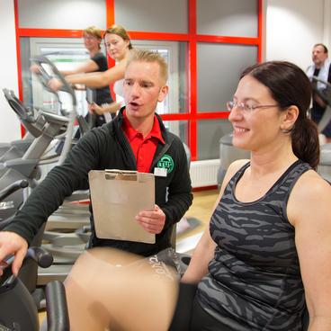 Trainer erkl&auml;rt einer Frau Einstellungen an einem Trainingsger&auml;t im Fitnessstudio, im Hintergrund weitere Personen beim Training.