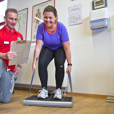 Ein Trainer notiert Daten, w&auml;hrend eine Frau auf einer Laufbandwaage steht und ihre Haltung &uuml;berpr&uuml;ft.