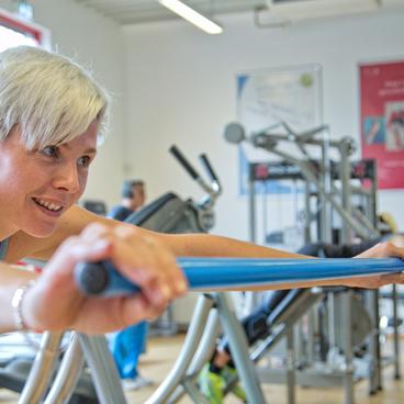 Frau mit kurzem grauem Haar trainiert mit einem blauen Widerstandsband in einem Fitnessstudio.