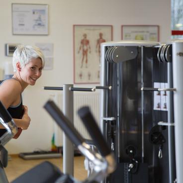 Frau mit kurzer blonder Frisur l&auml;chelt, w&auml;hrend sie an Fitnessger&auml;ten in einem modernen Fitnessstudio trainiert.