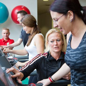 F&uuml;nf Personen trainieren an Laufb&auml;ndern in einem modernen Fitnessstudio mit bunten Gymnastikb&auml;llen im Hintergrund.