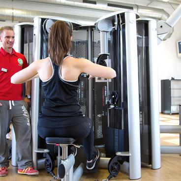 Zwei Frauen trainieren an Fitnessger&auml;ten in einem modernen Fitnessstudio, ein Trainer beobachtet sie.