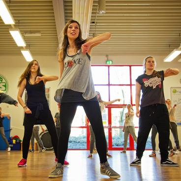 Gruppe junger Frauen tanzt gemeinsam in einem gro&szlig;en Raum mit Holzboden und gro&szlig;en Fenstern.