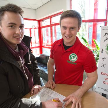 Zwei junge M&auml;nner stehen an einem Tisch, einer tr&auml;gt ein rotes T-Shirt mit Vereinslogo und hilft beim Ausf&uuml;llen eines Formulars.