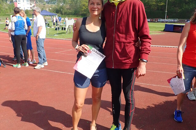 Zwei Personen posieren auf einer Laufbahn, eine h&auml;lt eine Urkunde, im Hintergrund sind weitere Teilnehmer sichtbar.