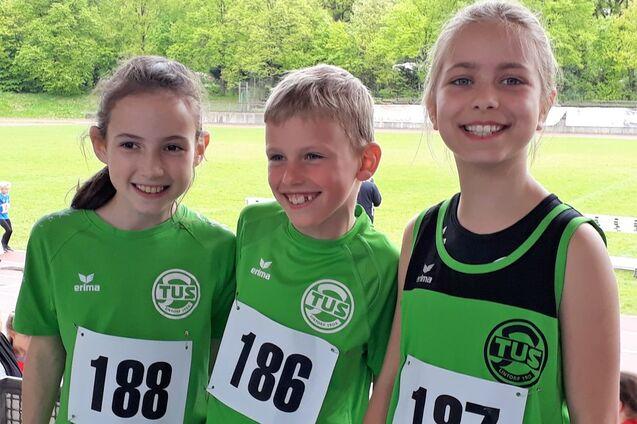 Drei Kinder in gr&uuml;nen Sporttrikots mit Startnummern l&auml;cheln in die Kamera vor einem Sportplatz.