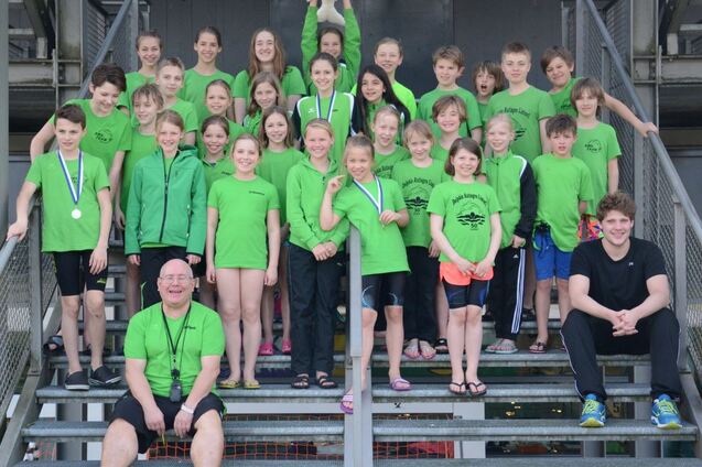 Gruppe von schwimmenden Kindern in gr&uuml;nen T-Shirts steht auf Treppen, einige halten Medaillen; ein Trainer sitzt vorne.