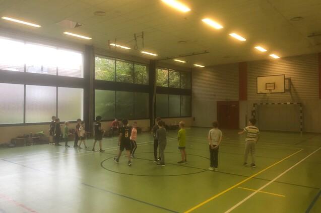 Gruppe von Kindern spielt in einer Sporthalle mit Basketballkorb, helle Fenster und gr&uuml;ne Bodenfl&auml;che.