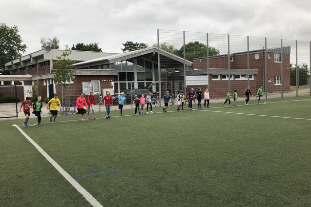 Gruppe von Kindern in Sportkleidung trainiert auf einem Kunstrasenfeld vor einem Sportgeb&auml;ude.