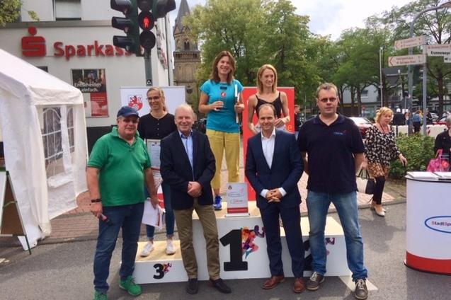 Gruppe von Personen steht auf einem Siegertreppchen vor einer Sparkassen-Filiale in einer Stadt, umgeben von Zuschauern.