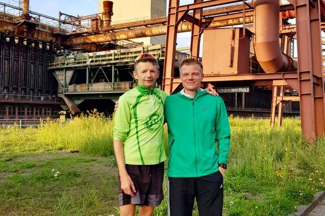 Zwei M&auml;nner in Sportbekleidung stehen vor der Kokerei Zollverein, umgeben von gr&uuml;ner Wiese und industrieller Architektur.