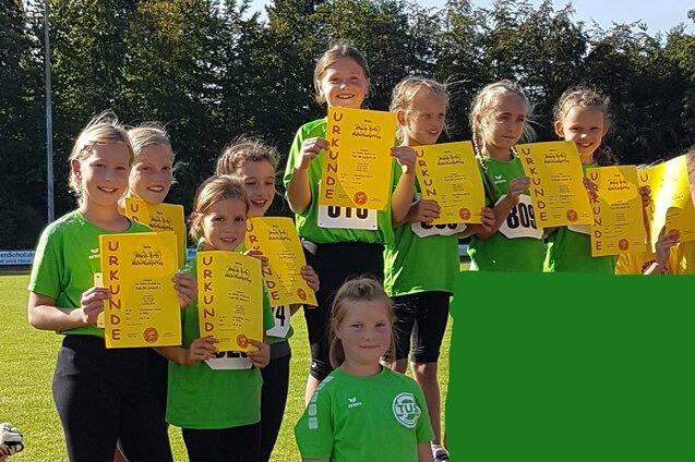 Gruppe von zehn M&auml;dchen in gr&uuml;nen T-Shirts, die stolz ihre Urkunden auf dem Siegerpodest zeigen.