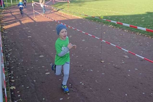 Ein Junge in gr&uuml;ner Sportkleidung l&auml;uft auf einer Laufbahn, umgeben von herbstlichen B&auml;umen und Zuschauern.