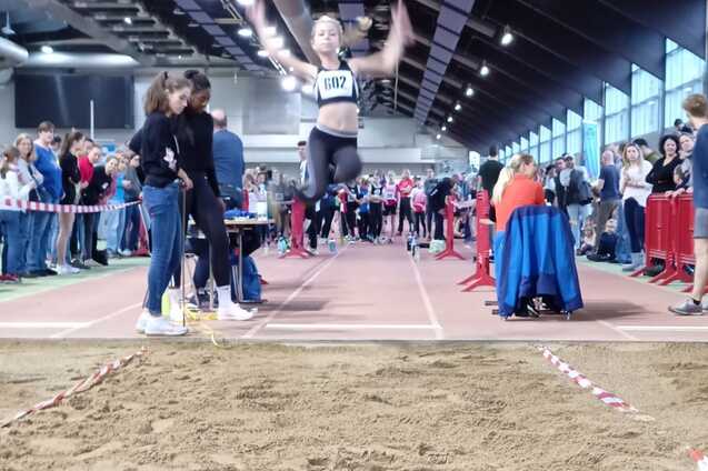 Sprinterin springt in einem Leichtathletikwettbewerb &uuml;ber die Sandgrube, Zuschauer und Teilnehmer im Hintergrund.