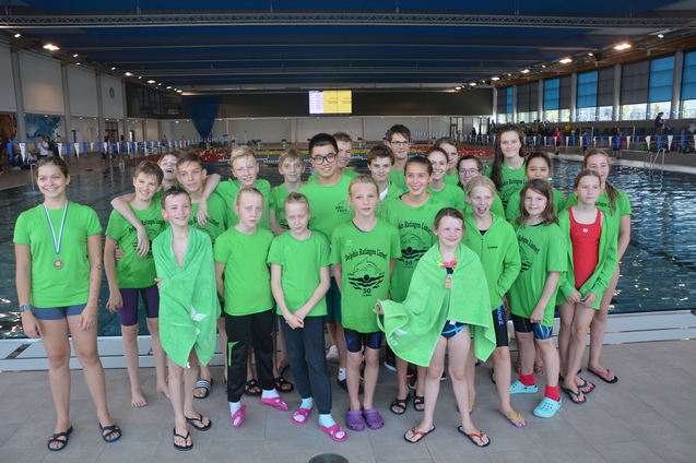 Gruppe von Schwimmern in gr&uuml;nen T-Shirts am Beckenrand eines Schwimmbades, l&auml;chelnd posierend f&uuml;r ein Foto.