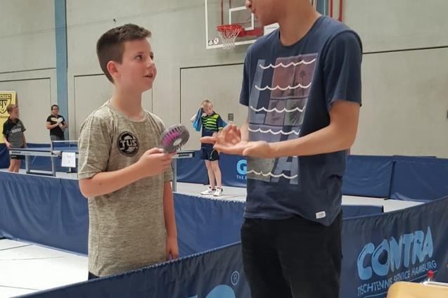 Zwei Jungen sprechen in einer Tischtennishalle, einer h&auml;lt einen Tischtennisschl&auml;ger, der andere zeigt Interesse.