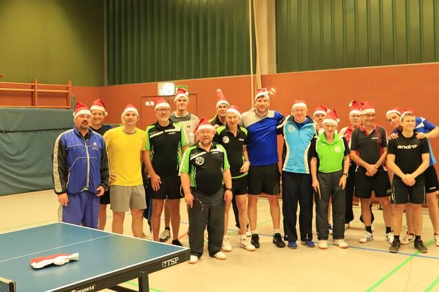 Gruppe von Tischtennisspielern in Weihnachtsm&uuml;tzen posiert gemeinsam in einer Sporthalle neben einem Tischtennistisch.