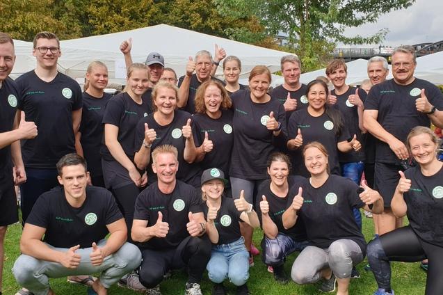 Gruppe von 16 Personen in schwarzen T-Shirts mit Daumen hoch, umgeben von Zelten und B&auml;umen im Freien.