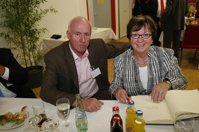 Zwei Personen sitzen an einem Tisch, l&auml;cheln und halten ein offenes Buch, umgeben von Getr&auml;nken und Essen.