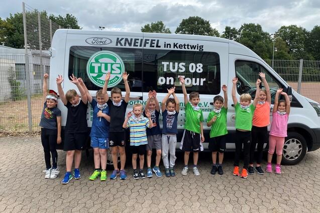 Gruppe von Kindern in Sportkleidung steht vor einem Vereinsbus mit "TUS 08 on tour" Aufdruck und hebt die H&auml;nde.