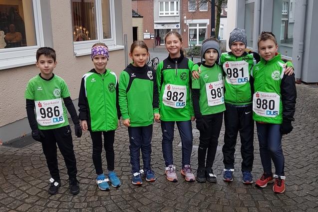 Gruppe von sieben Kindern in gr&uuml;nen Sportshirts und Startnummern auf einem Kopfsteinpflasterweg vor Geb&auml;uden.