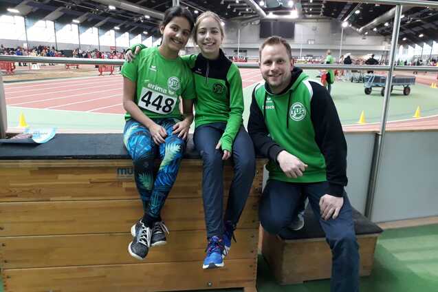 Drei Personen, zwei M&auml;dchen und ein Junge, in Sportoutfits sitzen auf einer Holzbank in einer Sporthalle.