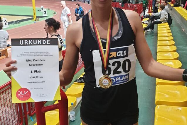 Sportlerin mit Medaille und Urkunden in der Hand, steht in einer Sporthalle mit gelben Sitzen im Hintergrund.
