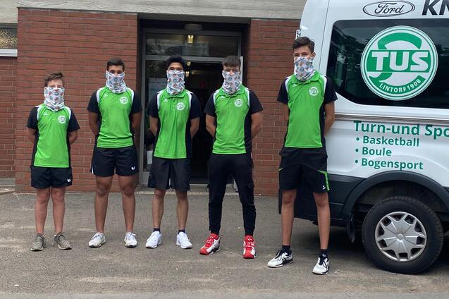 F&uuml;nf Sportler in gr&uuml;nen T-Shirts stehen vor einem Fahrzeug mit dem Logo des TUS, alle tragen Gesichtsmasken.