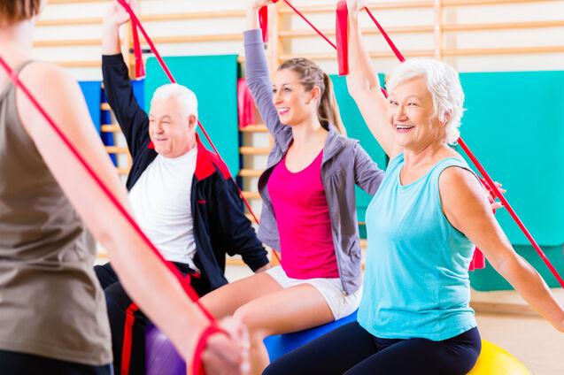 Gruppe von vier Personen, die in einem Fitnessraum mit Widerstandsb&auml;ndern &Uuml;bungen auf Gymnastikb&auml;llen ausf&uuml;hren.