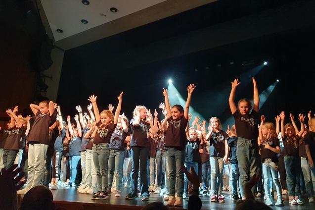 Gruppendarbiet von Kindern auf einer B&uuml;hne, die fr&ouml;hlich mit erhobenen H&auml;nden tanzen.