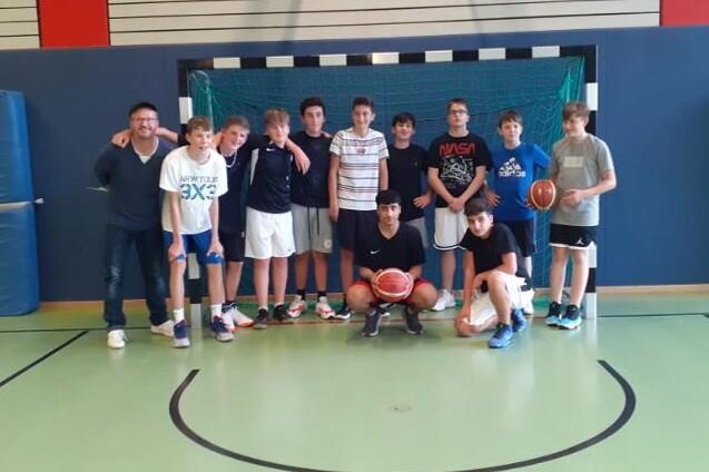 Gruppe von Jugendlichen mit Basketball im Sporthallen-Hintergrund, zwei Trainer stehen links neben den Spielern.