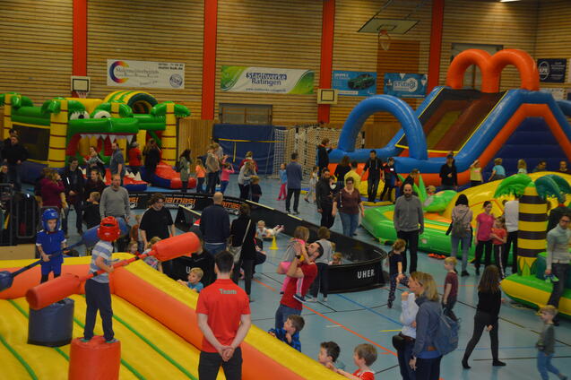 Bunte H&uuml;pfburgen und Spielstationen in einer Halle, zahlreiche Kinder und Erwachsene genie&szlig;en Aktivit&auml;ten und Spa&szlig;.