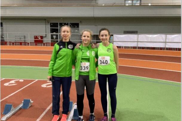Drei Sportlerinnen in gr&uuml;nen Trikots stehen l&auml;chelnd auf einer Leichtathletikanlage mit Startbl&ouml;cken.