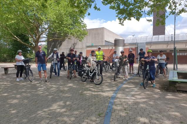 Gruppe von Menschen mit Fahrr&auml;dern auf einem Platz, umgeben von B&auml;umen und Geb&auml;uden. Einige tragen Helme.