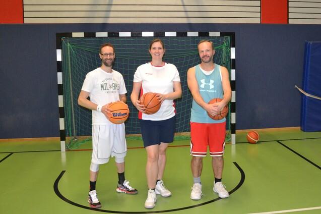 Drei Personen stehen mit Basketballs vor einem Basketballkorb in einer Sporthalle.