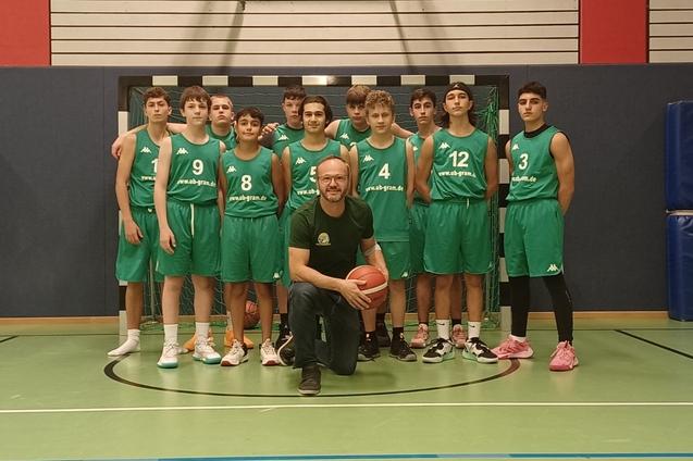 Basketballmannschaft in gr&uuml;nen Trikots posiert gemeinsam auf dem Spielfeld, ein Trainer h&auml;lt den Ball in der Mitte.