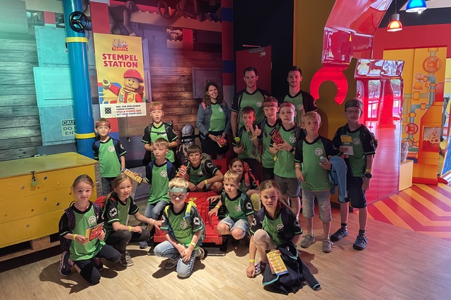 Gruppe von Kindern mit Betreuern in T-Shirts mit einer Stempelstation im Hintergrund in einem bunten Raum.