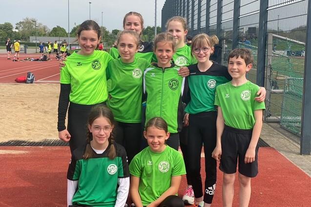 Gruppe von neun Kindern in gr&uuml;nen Sporttrikots, die auf einem Sportplatz l&auml;cheln und posieren.