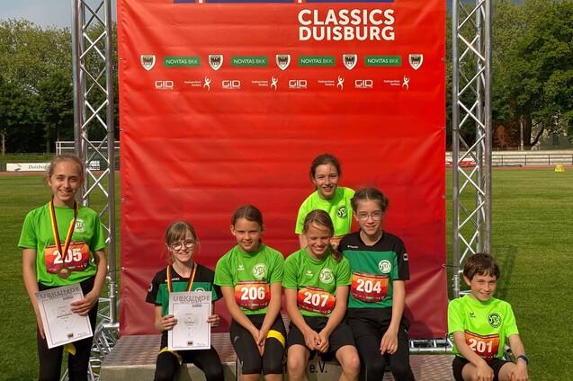 Gruppe von sieben Kindern in gr&uuml;nen Sportanz&uuml;gen, posierend vor einem roten Banner nach einem Laufwettbewerb.