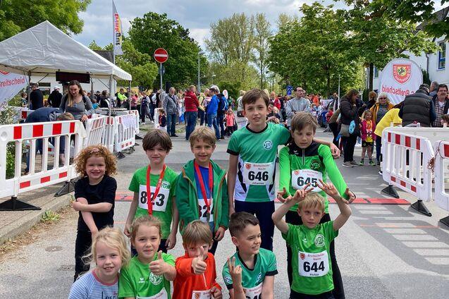 Gruppeneinstellung von sieben Kindern in Sportkleidung mit Medaillen nach einem Laufwettbewerb, im Hintergrund eine Startbanner.