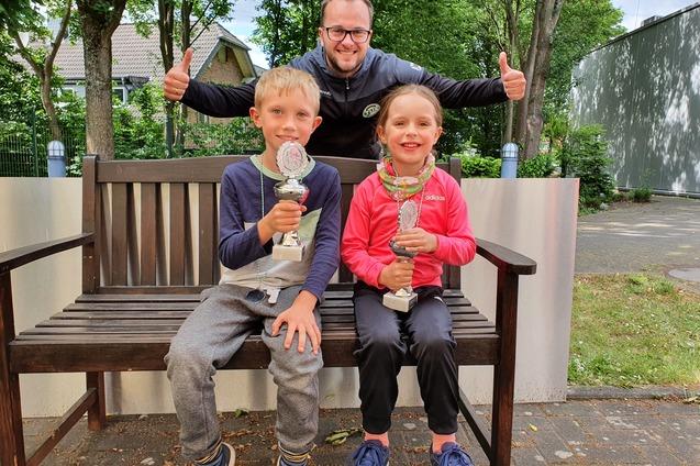 Zwei Kinder mit Pokalen sitzen auf einer Bank, w&auml;hrend ein l&auml;chelnder Erwachsener dahinter mit Daumen hoch steht.