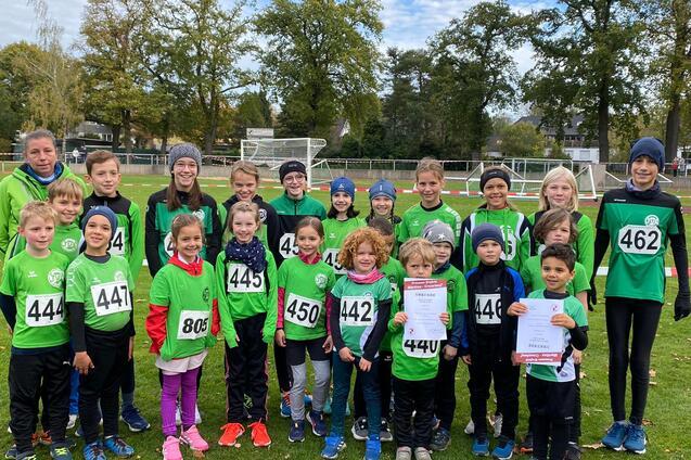 Gruppe von 25 Kindern in gr&uuml;nen Trikots mit Startnummern auf einem Sportplatz, einige halten Urkunden in den H&auml;nden.