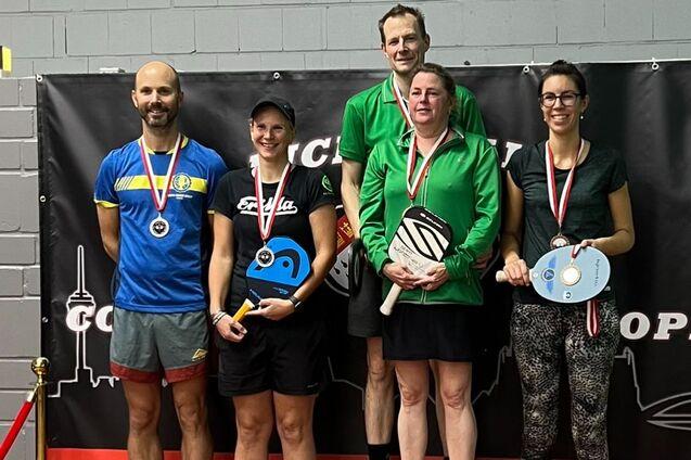 F&uuml;nf Personen stehen auf einem Siegerpodest mit Medaillen, umgeben von einer Tischtennisarena mit Werbebanner.