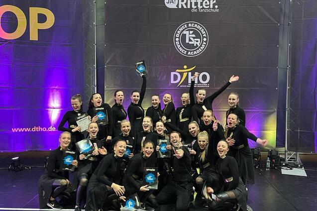 Gruppe von T&auml;nzerinnen in schwarzen Outfits h&auml;lt Pokale und jubelt auf einer B&uuml;hne mit buntem Hintergrund.