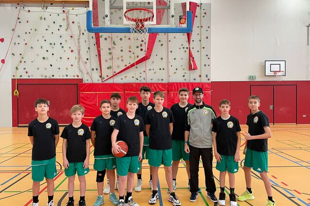 Gruppe von zw&ouml;lf Jugendlichen in Sportkleidung mit Basketball in einer Sporthalle vor einem Korb.